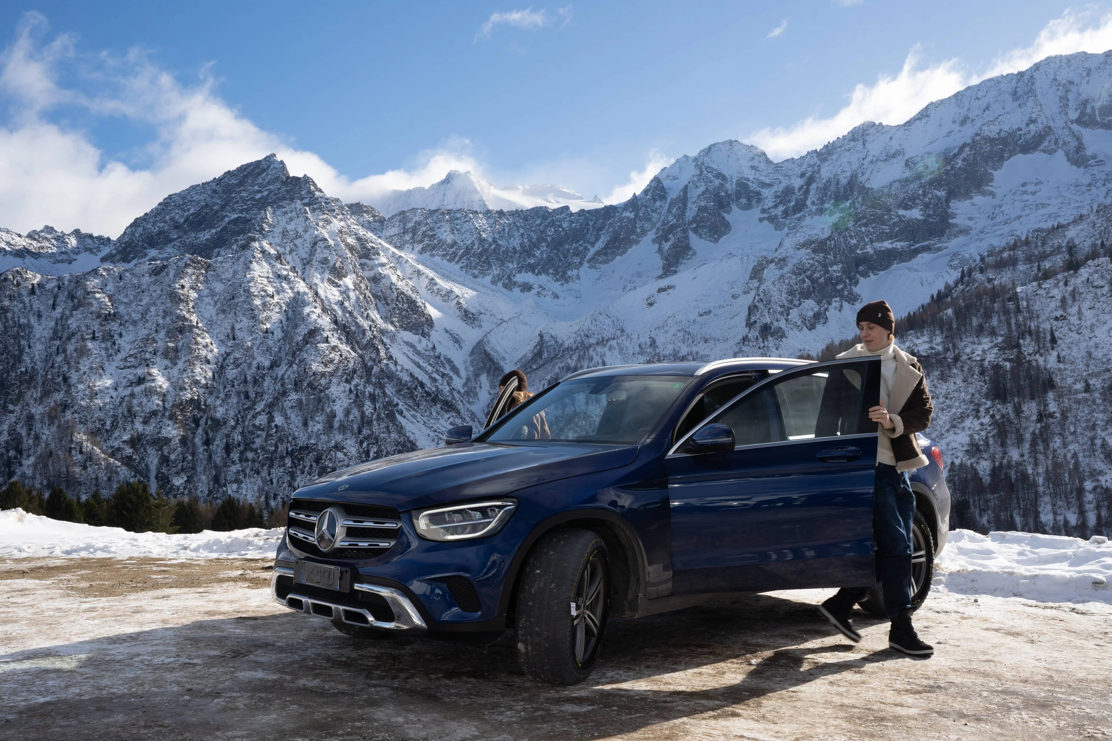 Person standing next to a blue Mercedes-Benz SUV in a snowy mountainous area with ARCOL snow socks installed